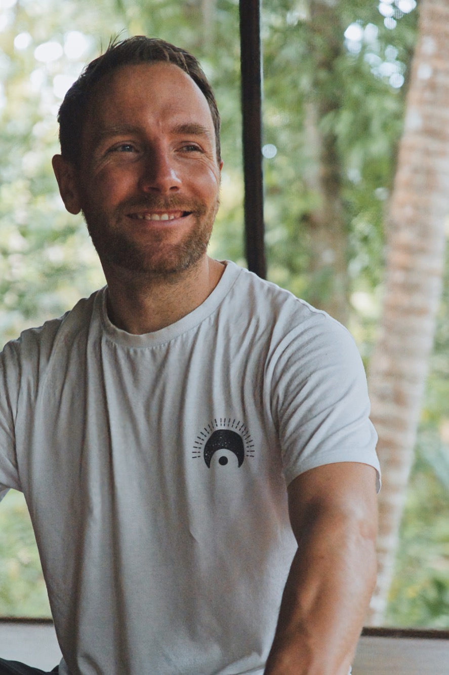 Arogyaspirit-Breathwork-Florian Ein Mann mit kurzen braunen Haaren und Bart sitzt lächelnd drinnen. Er trägt ein weißes T-Shirt mit einem kleinen kreisförmigen schwarzen Muster auf der Brust. Im Hintergrund sind grüne Bäume und Sonnenlicht zu sehen.